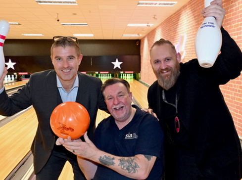 Three men at a bowling alley. Two standing holding skittles. One in wheelchair holding bowling ball. 